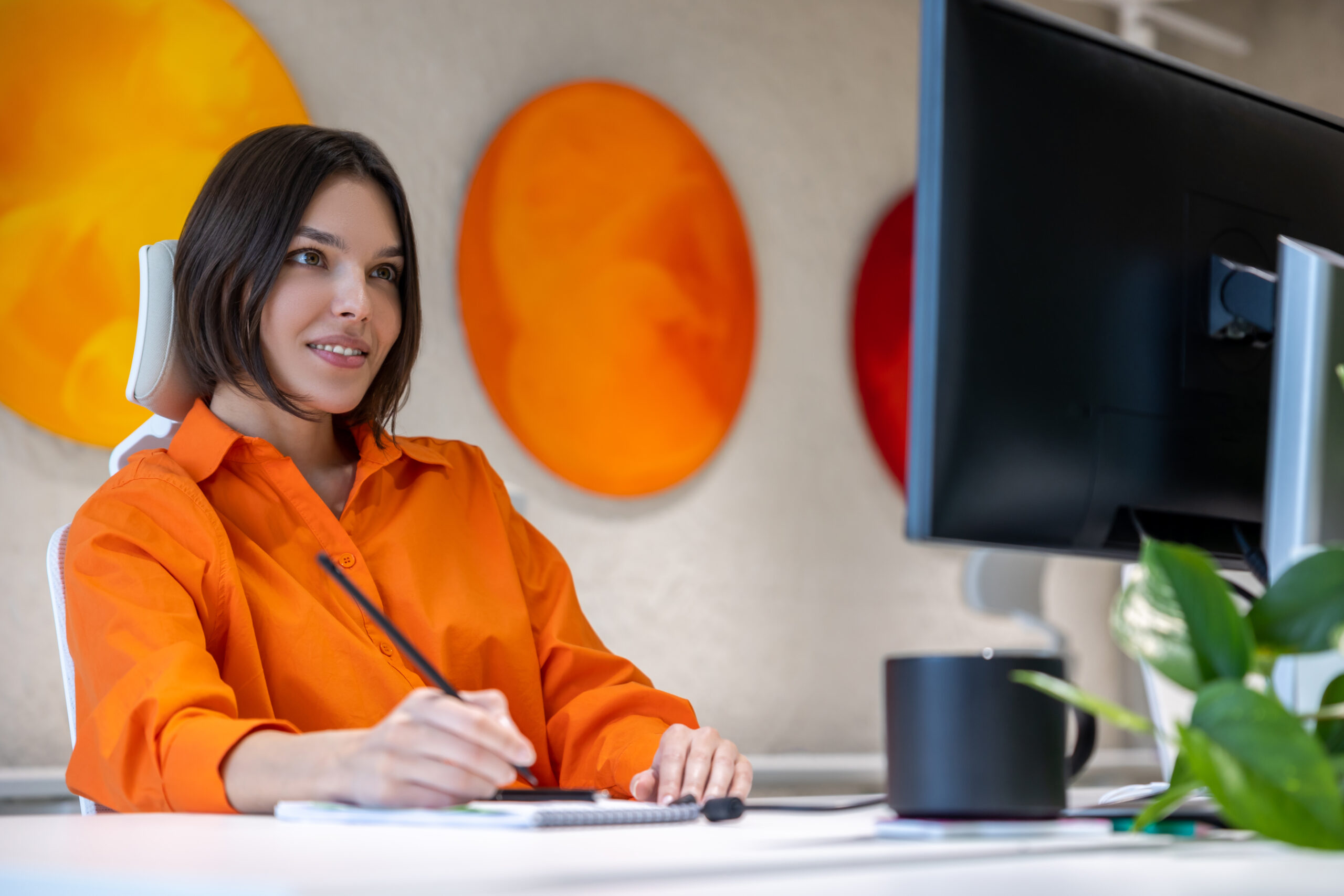 concentrated-company-employee-seated-office-desk-looking-computer-monitor-while-making-notes-her-notebook-scaled.jpg Woman working behind her desk